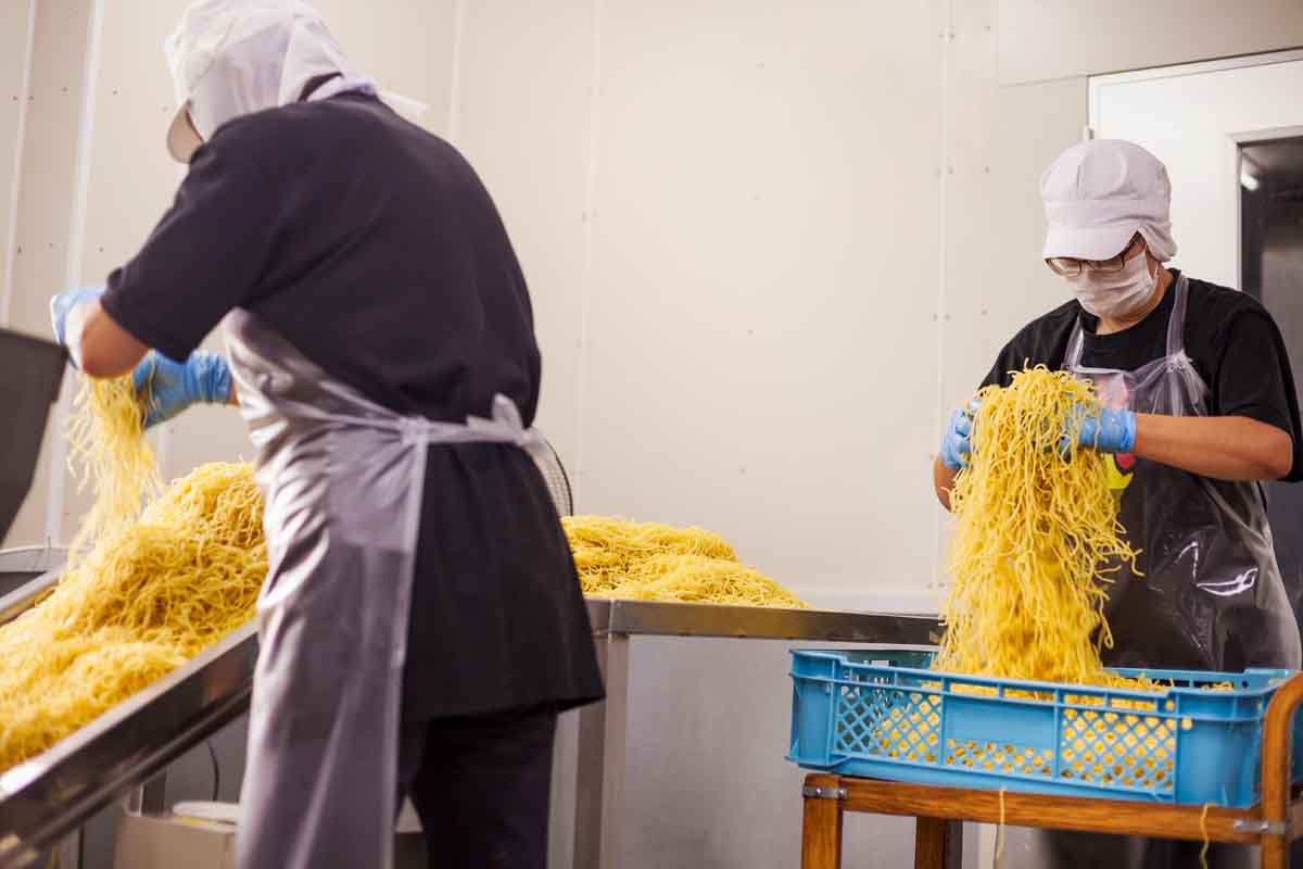 Workers in aprons and hats collecting freshly cut noodles from the conveyor belt to package and sell.